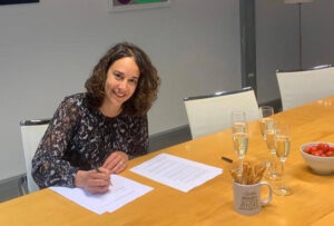 Heleen Blesgraaf tekent een contract bij Uitgeverij Moon. Op de houten tafel staan vijf glazen champagne, kaasstengels en aarbeiden.